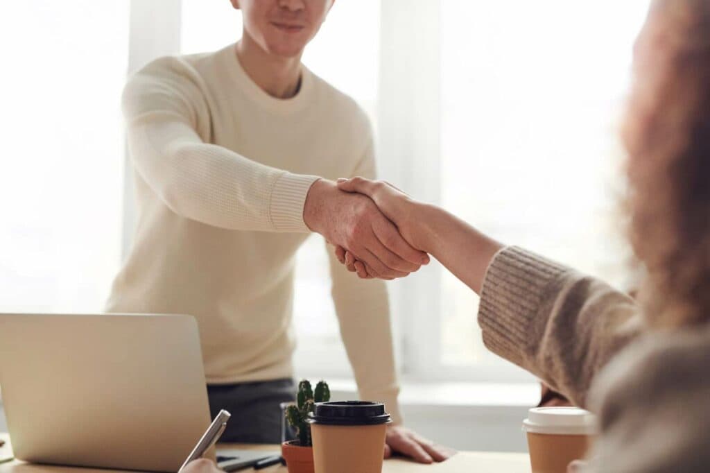 Two people shaking hands over a desk that had a to-go coffee cup and laptop